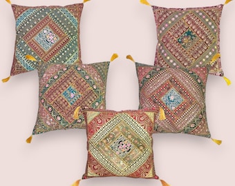 Puede incluir: Cinco cojines decorativos con intrincados patrones bordados en tonos rojos, verdes y dorados. Cada cojín presenta un diseño de diamante central y está adornado con una borla amarilla en cada esquina. Los cojines están dispuestos sobre un fondo rosa claro.