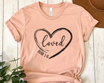 May include: A light pink t-shirt with a black heart graphic and the text "loved" inside the heart. Below the heart is the text "JOHN 3:16".