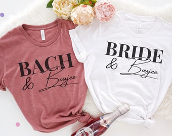 May include: Two white t-shirts with black text. The shirt on the left says "Bach & Boujee" in black text on a mauve shirt. The shirt on the right says "Bride & Boujee" in black text on a white shirt. A bottle of sparkling wine is in the center of the image.
