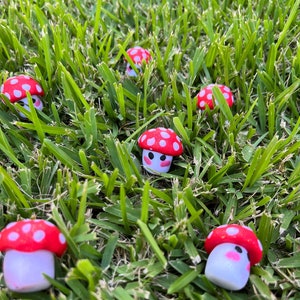 May include: A collection of miniature mushroom figurines with red and white spotted caps and white bodies, nestled amongst green grass. The mushrooms feature smiling faces and pink cheeks, creating a charming, playful scene.