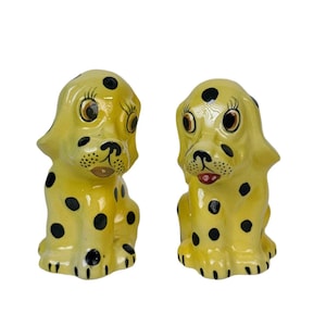 May include: A pair of ceramic salt and pepper shakers shaped like yellow puppies with black spots. The puppies have large, expressive eyes, black noses, and red mouths. They are sitting upright.