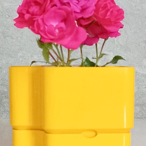 Puede incluir: Un vibrante arreglo de rosas rosas en una maceta amarilla brillante de forma única. Las flores tienen múltiples capas de pétalos y tallos verdes. La maceta tiene un diseño distintivo con una pequeña hendidura curva en la parte delantera.