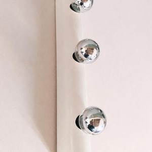 May include: A white wall-mounted light fixture with three chrome globe lights. The fixture has a sleek, minimalist design and is mounted on a light-colored wall. The light bulbs are reflective and shiny.