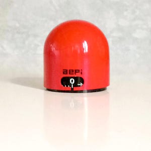 May include: A red kitchen timer with a black dial and the word "aepi" printed on the front. The dial is set to zero.