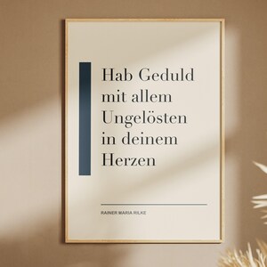 Könnte beinhalten: Ein rechteckiges Wandbild in einem hellen Holzrahmen mit cremefarbenem Hintergrund, schwarzem Text Hab Geduld mit allem Ungelösten in deinem Herzen, einem dunkelblauen vertikalen Balken auf der linken Seite und dem Namen Rainer Maria Rilke.