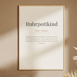 Può includere: Una stampa artistica da parete rettangolare in una cornice di legno chiaro con la parola tedesca Ruhrpottkind in carattere serif nero. Sotto la parola si trovano la pronuncia fonetica Meik e la classificazione del sostantivo, seguite da un testo.