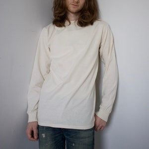 Puede incluir: Una persona con una camisa blanca de manga larga y pantalones vaqueros azules. La camisa tiene cuello redondo y mangas largas.