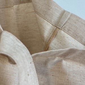 May include: Close-up of a beige tote bag made of a textured fabric. The bag's interior and handles are visible, showcasing the stitching and construction details. The neutral color and simple design suggest a reusable shopping bag or everyday carryall.