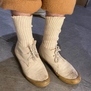 May include: A pair of off-white suede lace-up shoes with tan soles. The shoes are worn with cream-colored ribbed socks and tan-colored pants. The shoes appear to be well-worn.