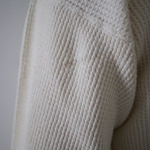 K&ouml;nnte beinhalten: Nahaufnahme eines wei&szlig;en Waffelstrickpullovers. Der Stoff ist weich und texturiert.