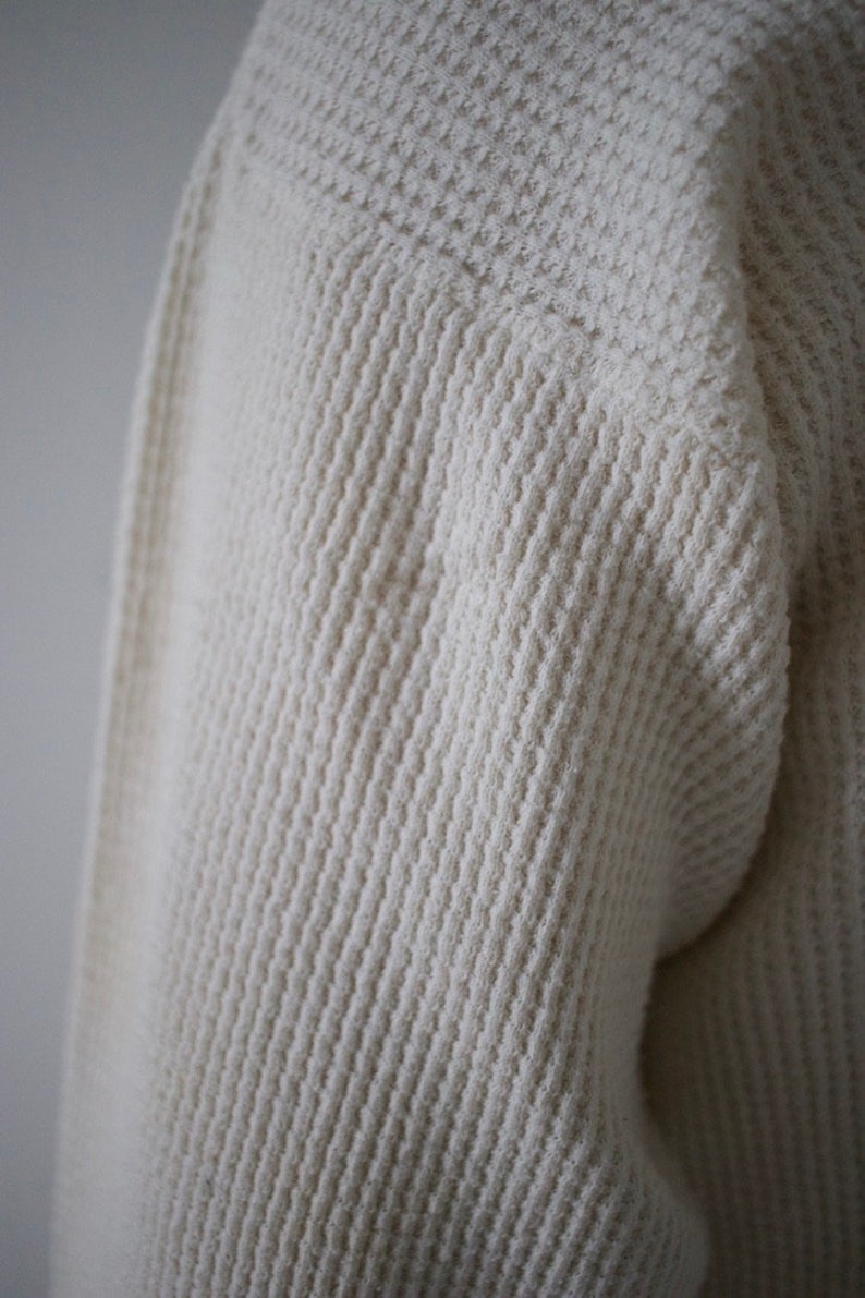 K&ouml;nnte beinhalten: Nahaufnahme eines wei&szlig;en Waffelstrickpullovers. Der Stoff ist weich und texturiert.