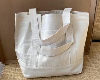 May include: A beige canvas tote bag with a rectangular shape and a front pocket. The bag has two handles and is sitting on a woven mat. The bag is made of a sturdy material.