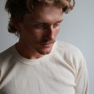 K&ouml;nnte beinhalten: Nahaufnahme einer Person, die ein cremefarbenes Langarmshirt aus Waffelpiqu&eacute; tr&auml;gt. Die Person hat hellbraunes Haar und schaut nach unten.