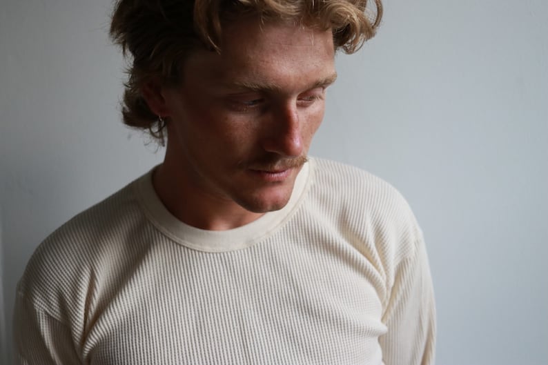 K&ouml;nnte beinhalten: Nahaufnahme einer Person, die ein cremefarbenes Langarmshirt aus Waffelpiqu&eacute; tr&auml;gt. Die Person hat hellbraunes Haar und schaut nach unten.