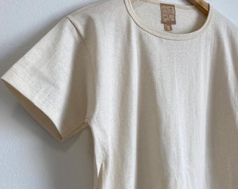 Könnte beinhalten: Ein schlichtes, cremefarbenes T-Shirt mit kurzen Ärmeln hängt an einem Holzbügel. Das T-Shirt hat einen Rundhalsausschnitt und ein kleines braunes Etikett mit dem Text "Organic Cotton" und "Made in Japan" darauf.