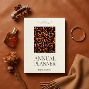 May include: An "Annual Planner" book with a leopard print design on the cover, alongside a gold bracelet, perfume bottle, hair clip, dried rose, and a tan scarf. The book is part of "A Wardrobe Logic Collection" by @lawton_home.