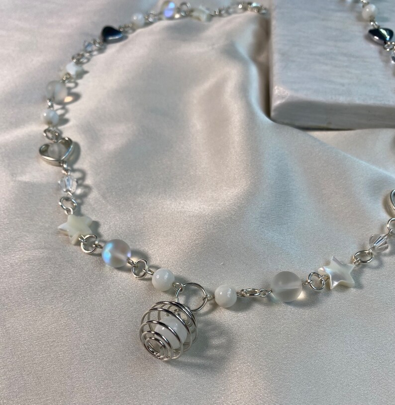 Puede incluir: Un delicado collar plateado con abalorios de estrellas, corazones y cuentas iridiscentes. Un colgante de orbe envuelto en alambre cuelga del centro. El collar se exhibe sobre una superficie de sat&eacute;n blanco.