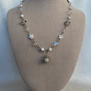 Puede incluir: Un delicado collar plateado con un colgante con un orbe blanco encerrado en una jaula de alambre. El collar incluye dijes blancos en forma de estrella y coraz&oacute;n, cuentas iridiscentes y cuentas redondas blancas. El collar se exhibe en un maniqu&iacute; de tono neutro.
