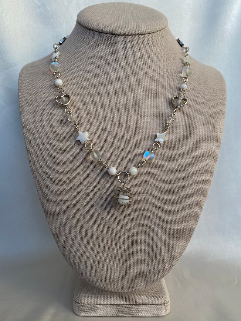 Puede incluir: Un delicado collar plateado con un colgante con un orbe blanco encerrado en una jaula de alambre. El collar incluye dijes blancos en forma de estrella y coraz&oacute;n, cuentas iridiscentes y cuentas redondas blancas. El collar se exhibe en un maniqu&iacute; de tono neutro.