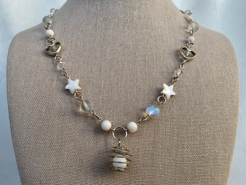 Puede incluir: Collar plateado con una variedad de cuentas, incluyendo esferas blancas, cristales transparentes y orbes iridiscentes. El collar tambi&eacute;n incluye colgantes en forma de estrella y coraz&oacute;n, y un colgante con alambre y una cuenta blanca.