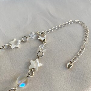 Puede incluir: Un collar plateado con una cadena fina, adornado con abalorios en forma de estrella blanca, cuentas de cristal transparente y una perla iridiscente. El collar incluye un dije en forma de coraz&oacute;n en el cierre.