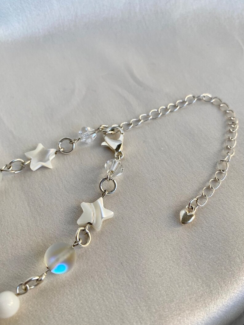 Puede incluir: Un collar plateado con una cadena fina, adornado con abalorios en forma de estrella blanca, cuentas de cristal transparente y una perla iridiscente. El collar incluye un dije en forma de coraz&oacute;n en el cierre.
