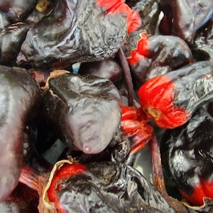 May include: A collection of dried chili peppers with wrinkled, dark purple-black skins and vibrant red sections visible at the base and stem ends. The peppers have a bulbous, irregular shape and a glossy, dehydrated texture throughout the pile.