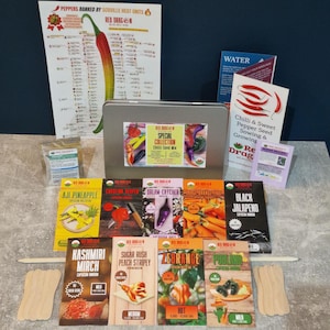 May include: A Red Dragon brand chili seed collection kit featuring a silver metal tin, multiple seed packets labeled with varieties like Aji Pineapple and Carolina Reaper, a Scoville heat unit chart, a growing guide, and wooden plant markers.