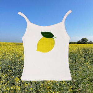 May include: White camisole top with thin straps, featuring a yellow lemon applique with a green leaf. The lemon design is centered on the front of the top. The background shows a field of yellow flowers under a blue sky.