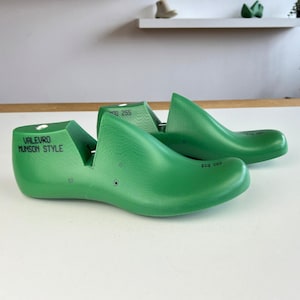 May include: Two bright green shoe lasts, with the text "VALEURO MUNSON STYLE" on one. The lasts are made of a solid material and are designed to shape shoes. The background is a white surface with a shelf and plants.