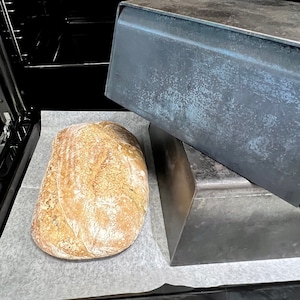 Op de afbeelding: Een versgebakken brood op bakpapier in een oven. Een donkere metalen broodpan is boven het brood geplaatst. Het brood heeft een goudbruine korst en is klaar om van te genieten.