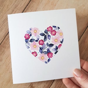 May include: A white card with a pink and purple floral heart design. The heart is made of pink and purple flowers with blue leaves and white dots.