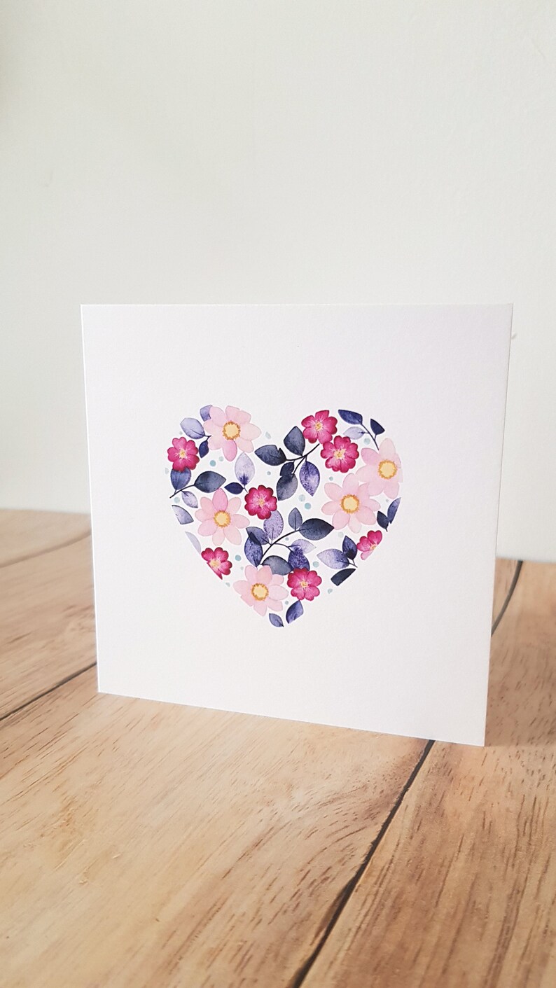May include: A white card with a pink and purple floral heart design. The heart is made up of pink and purple flowers with green leaves and blue dots.