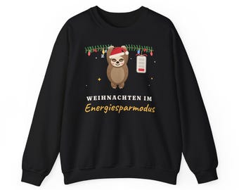 May include: A black sweatshirt featuring a graphic of a sloth wearing a Santa hat, hanging from a green pine branch with colorful Christmas lights. Next to the sloth is a low battery icon. The text reads WEIHNACHTEN IM Energiesparmodus.