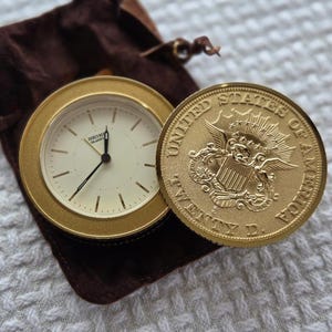 May include: A gold-toned Seiko Quartz clock with a cream-colored face and black hands, next to a gold coin with the words "UNITED STATES OF AMERICA" and an eagle emblem. The clock and coin rest on a brown velvet pouch.