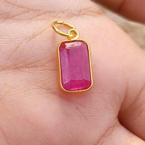 May include: A rectangular, ruby-colored gemstone pendant with a gold-colored bezel and bail. The pendant is a vibrant pinkish-red hue, and the gold setting provides a contrasting frame. The jewelry piece is likely a necklace charm or earring component.