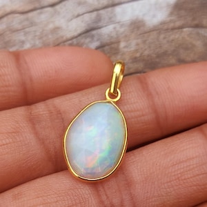 Puede incluir: Un colgante de oro con una piedra preciosa de ópalo ovalada. El ópalo muestra un juego de colores con destellos iridiscentes de verde, azul y rosa. El colgante tiene un diseño sencillo y elegante, adecuado para el uso diario.