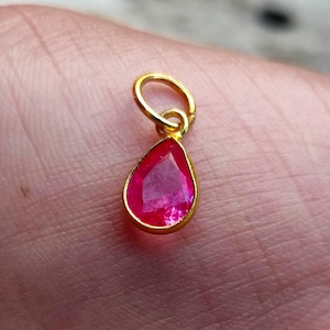 Puede incluir: Un colgante dorado con una gema rosa brillante en forma de lágrima. La gema está engastada en un bisel dorado y tiene un pequeño lazo dorado en la parte superior para una cadena. El colgante se muestra sobre un fondo color piel.