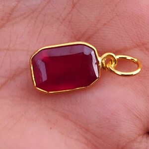 Puede incluir: Un colgante rectangular de piedra preciosa rojo oscuro con un bisel y un lazo dorados. El colgante se sostiene en una mano, mostrando el color y el diseño. Esta pieza de joyería es adecuada para collares o pulseras.