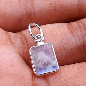 Puede incluir: Un colgante rectangular con un marco plateado y una piedra azul claro y blanca. El colgante tiene un pequeño lazo en la parte superior para una cadena. La piedra tiene un aspecto nublado.