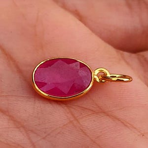 May include: A gold pendant with a large, oval-shaped, faceted ruby gemstone. The ruby is a deep pink color and is set in a gold bezel. The pendant has a small gold loop for attaching to a chain.