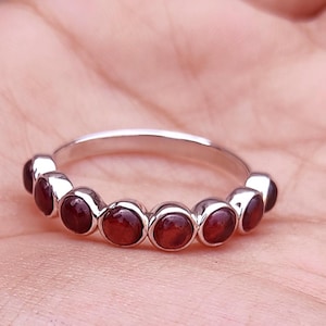 Könnte beinhalten: Ein silberner Ring mit einer Reihe von runden, dunkelroten Edelsteinen. Der Ring wird in einer Hand gehalten, was das polierte Metall und die leuchtende Farbe der Steine zeigt. Das Design des Rings ist schlicht und elegant. Die Edelsteine sind in einer Reihe angeordnet.