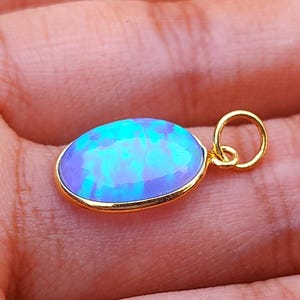 Op de afbeelding: Een gouden hanger met een ovale, iriserende opaalsteen. De opaal toont tinten blauw en groen. De hanger wordt in een hand gehouden, wat de grootte en het ontwerp van het sieraad benadrukt. De opaal is gezet in een gouden rand.