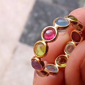 May include: A gold band ring featuring a series of small, round, cabochon-cut gemstones in various colours including pink, light blue, lime green, purple, and olive green, set in individual circular gold bezels arranged in a continuous circular pattern.