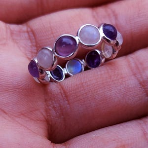 Puede incluir: Un anillo de plata con una serie de piedras preciosas redondas en tonos morados, lavanda y blancos. El anillo está engastado con múltiples piedras, creando una banda continua de color. El anillo se muestra sobre un fondo rosado.