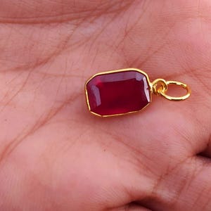 Könnte beinhalten: Ein rechteckiger Anhänger mit tiefrotem Edelstein, goldfarbener Lünette und Schlaufe. Der Anhänger wird in einer Hand gehalten, wodurch die satte Farbe und das Design des Schmuckstücks zur Geltung kommen. Die Goldeinfassung verstärkt den rubinartigen Stein.
