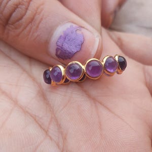 Può includere: Un anello d'oro con sei pietre rotonde di colore viola. L'anello è su una mano.