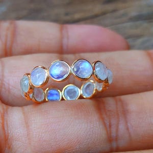 Peut inclure: Une bague en or ornée d'une série de pierres de lune rondes et iridescentes. Les pierres de lune sont de couleur bleu pâle et blanche, serties dans des chatons individuels en or. La bague est une bande continue.