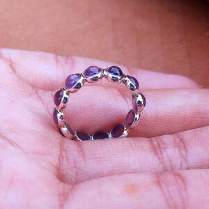 May include: A silver ring with a continuous band of oval, deep purple gemstones. The ring is set on a hand, showcasing the polished metal and the vibrant color of the stones. The ring is a piece of jewelry.