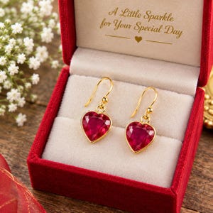 Könnte beinhalten: Ein Paar goldfarbene Ohrhänger mit facettierten herzförmigen roten Steinen, präsentiert in einer roten Samtschachtel mit dem Text A Little Sparkle for Your Special Day, der in Gold auf dem Innendeckel aufgedruckt ist, inklusive Herzsymbol.
